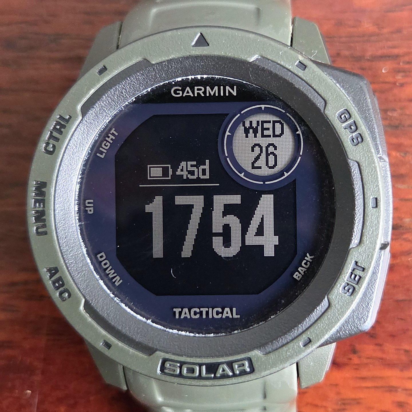 A Garmin Instinct Tactical with a sage green case and strap, its monochrome screen showing the time and a battery gauge.